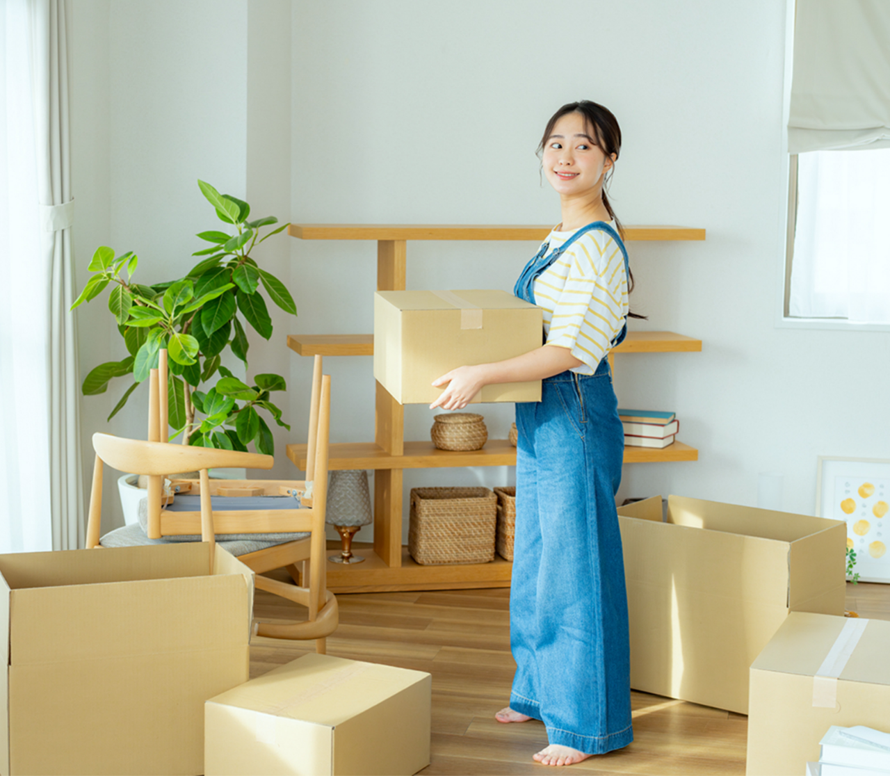 室内で段ボール箱を運ぶ女性が笑顔でカメラを見ている様子